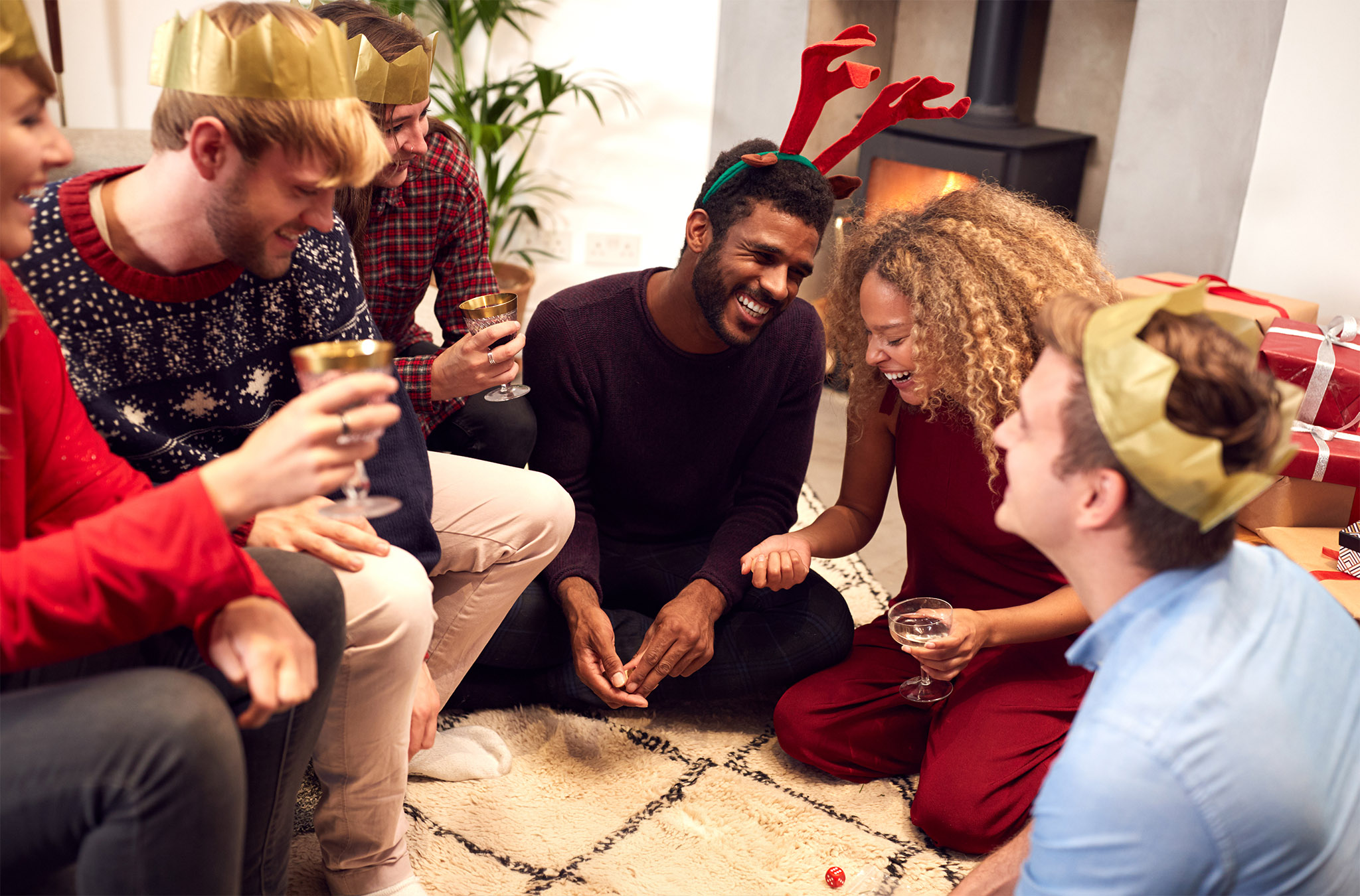 Friends wearing Christmas party hats and reindeer antlers laughing together while playing a holiday dice game on the floor during a festive gathering.